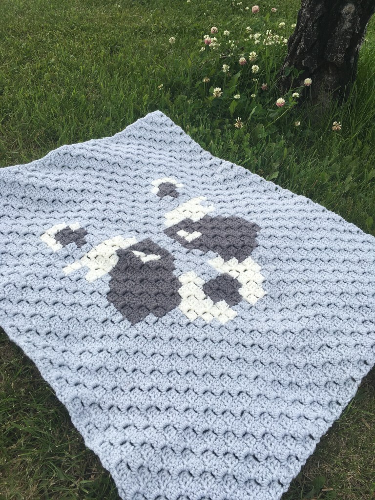 The Bandit stroller blanket lays spread on the grass. It is shades of grey and white with a raccoon face in the middle.